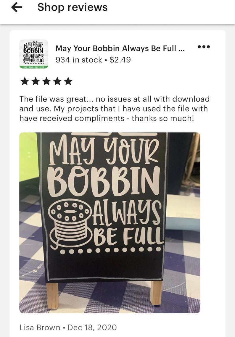 以下が含まれることがあります： 「May Your Bobbin Always Be Full」と白文字で書かれた黒い看板。看板には糸巻きが白くシルエットで描かれている。看板は木製のスタンドに置かれ、青と白の市松模様の表面の上に置かれている。