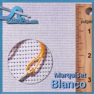 May include: Close-up of white cross-stitch fabric with a needle and yellow thread. A wooden ruler shows inches. The text "Marquiset Blanco" is visible.