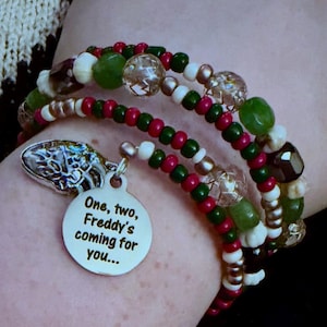 Op de afbeelding: Een armband met meerdere strengen kralen in groen, rood, wit en metallic. Er hangt een zilverkleurige metalen bedel aan met de tekst: One, two, Freddy's coming for you... Ook is er een tweede zilverkleurige bedel in de vorm van een klauw.