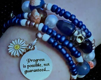 Op de afbeelding: Een armband met meerdere strengen met blauwe, witte en parel kralen. Een zilveren bedel met de tekst "Progress is possible, not guaranteed..." hangt naast een madeliefjesbedel. De armband heeft een mix van ronde en ovale kralen.