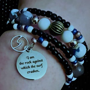 Könnte beinhalten: Mehrreihiges Armband mit schwarzen, weißen, blauen und goldenen Perlen. Ein silberner Anhänger mit Wellenmuster und der Aufschrift "I am the rock against which the surf crashes" hängt daran. Das Armband wird am Handgelenk getragen.