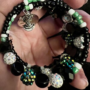 May include: A black and green beaded bracelet with a silver charm. The bracelet is made with black, white, and green beads, and has a silver charm in the shape of a snake.