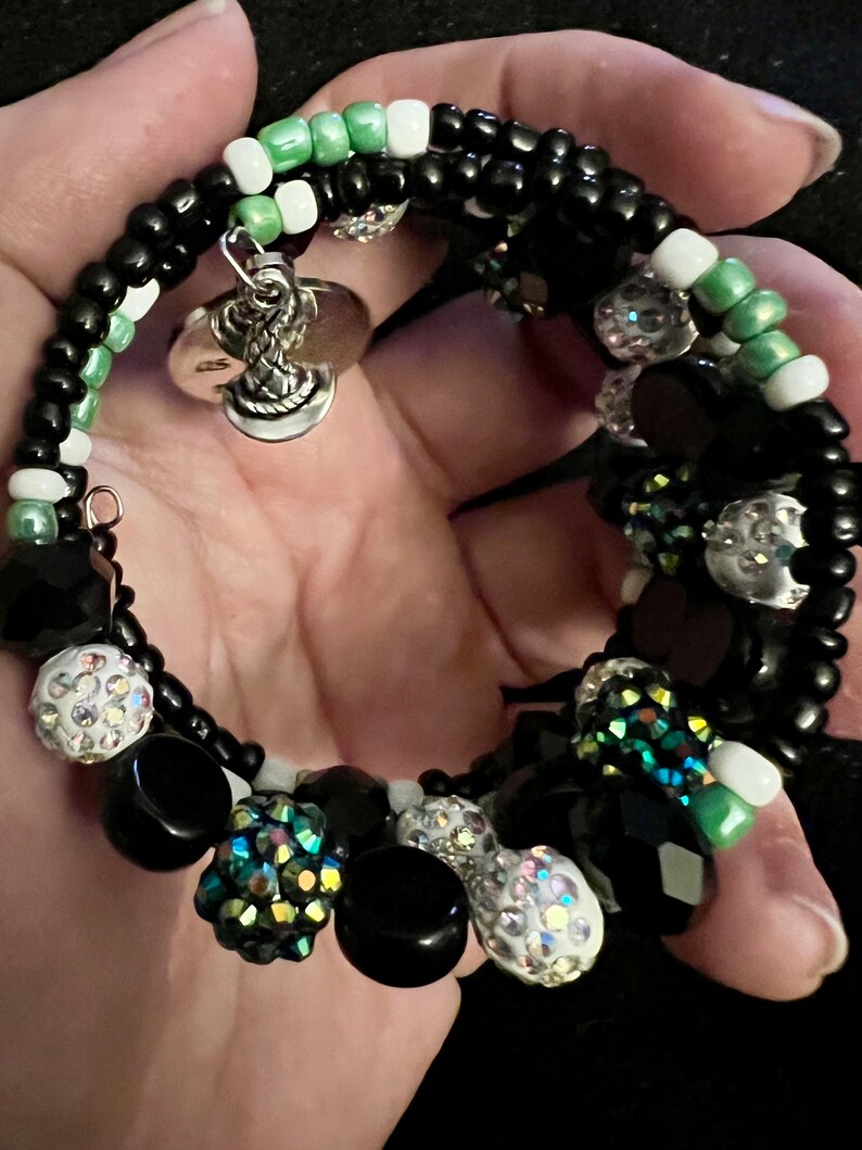 May include: A black and green beaded bracelet with a silver charm. The bracelet is made with black, white, and green beads, and has a silver charm in the shape of a snake.
