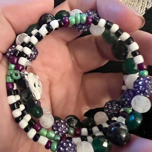 May include: A multi-strand beaded bracelet with black, white, green, purple, and white beads. The bracelet features a variety of bead shapes and sizes, including round, square, and irregular.
