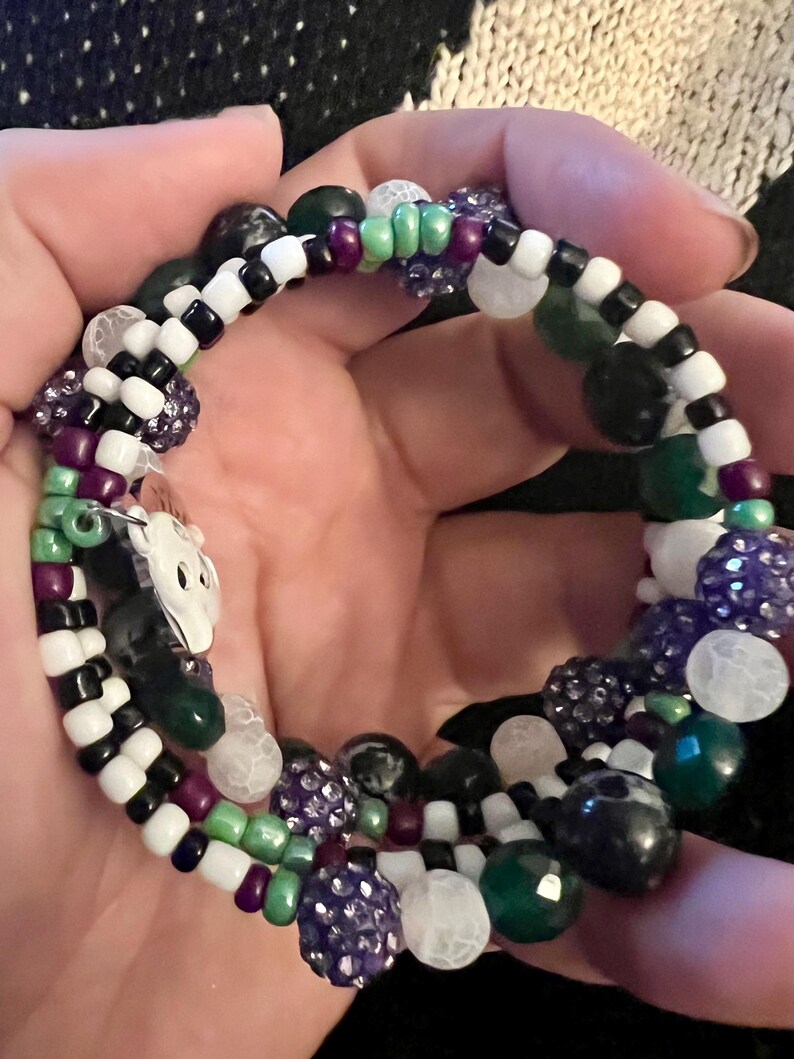 May include: A multi-strand beaded bracelet with black, white, green, purple, and white beads. The bracelet features a variety of bead shapes and sizes, including round, square, and irregular.