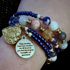 May include: A multi-strand beaded bracelet featuring dark blue, white, and translucent stone beads accented with gold-toned spacers. The bracelet includes a gold-toned heart charm and a round gold-toned pendant engraved with the text: Keep marching 'cause your ancestors are all the proof you need that progress is possible, not guaranteed.
