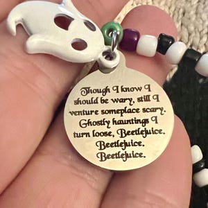 May include: A silver charm with a white ghost charm attached to a beaded bracelet. The charm has an inscription that reads "Though I know I should be wary, still I venture someplace scary. Ghostly hauntings I turn loose, Beetlejuice. Beetlejuice. Beetlejuice."