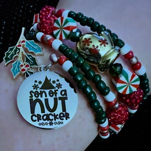 Op de afbeelding: Een stapel armbanden met kerstthema. De armbanden hebben groene, rode en witte kralen, samen met bedels zoals een gouden bel, hulstbladeren en een ronde bedel met de tekst "son of a nutcracker". De armbanden hebben ook snoep- en glitteraccenten.