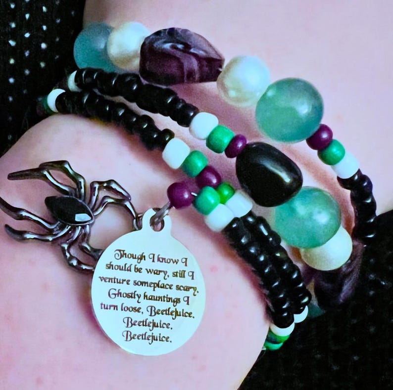May include: A black and white beaded bracelet with a silver spider charm and a silver tag that reads "Though I know I should be wary, still I venture someplace scary. Ghostly hauntings I turn loose, Beetlejuice. Beetlejuice. Beetlejuice."