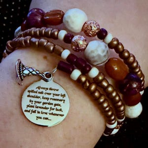 May include: A multi-strand beaded bracelet featuring metallic bronze, white, dark purple, and amber-colored beads. A silver-toned circular charm hangs from the bracelet, engraved with the text: Always throw spilled salt over your left shoulder, keep rosemary by your garden gate, plant lavender for luck, and fall in love whenever you can...