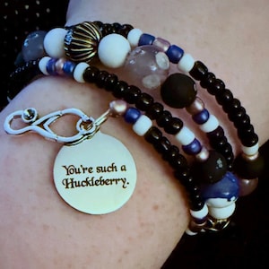 Könnte beinhalten: Ein mehrreihiges Perlenarmband mit schwarzen, weißen, blauen und metallischen Perlen. Ein silberfarbener Stethoskop-Anhänger ist an einem runden Metallanhänger befestigt, der mit dem Text "You're such a Huckleberry" graviert ist. Das Armband ist zum Wickeln um das Handgelenk gedacht.