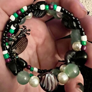 May include: A multi-strand bracelet with black, white, green, and purple beads. The bracelet features a variety of charms, including a heart-shaped charm with a design, a ghost charm, and a black and white charm.