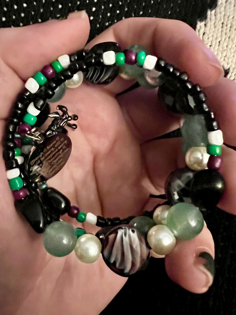 May include: A multi-strand bracelet with black, white, green, and purple beads. The bracelet features a variety of charms, including a heart-shaped charm with a design, a ghost charm, and a black and white charm.