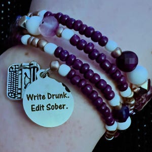 Könnte beinhalten: Mehrsträngiges Armband mit lila, weißen und goldenen Perlen. Ein silberner Anhänger mit dem Text "Write Drunk. Edit Sober." hängt am Armband. Das Armband wird am Handgelenk getragen.