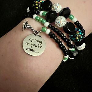May include: A multi-strand bracelet with black, white and green beads. The bracelet has a silver charm with the inscription "As long as you're mine..."