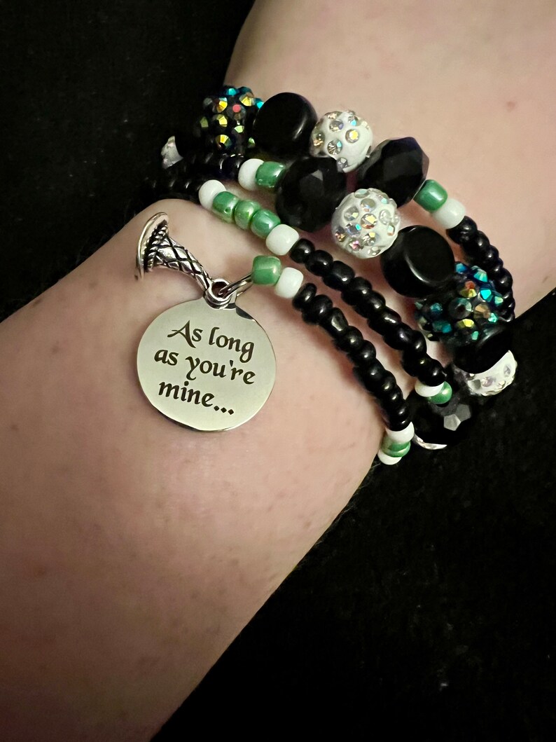 May include: A multi-strand bracelet with black, white and green beads. The bracelet has a silver charm with the inscription "As long as you're mine..."