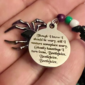 May include: A silver charm with a black spider design and a quote from the movie Beetlejuice. The quote reads: "Though I know I should be wary, still I venture someplace scary. Ghostly hauntings I turn loose, Beetlejuice. Beetlejuice. Beetlejuice."