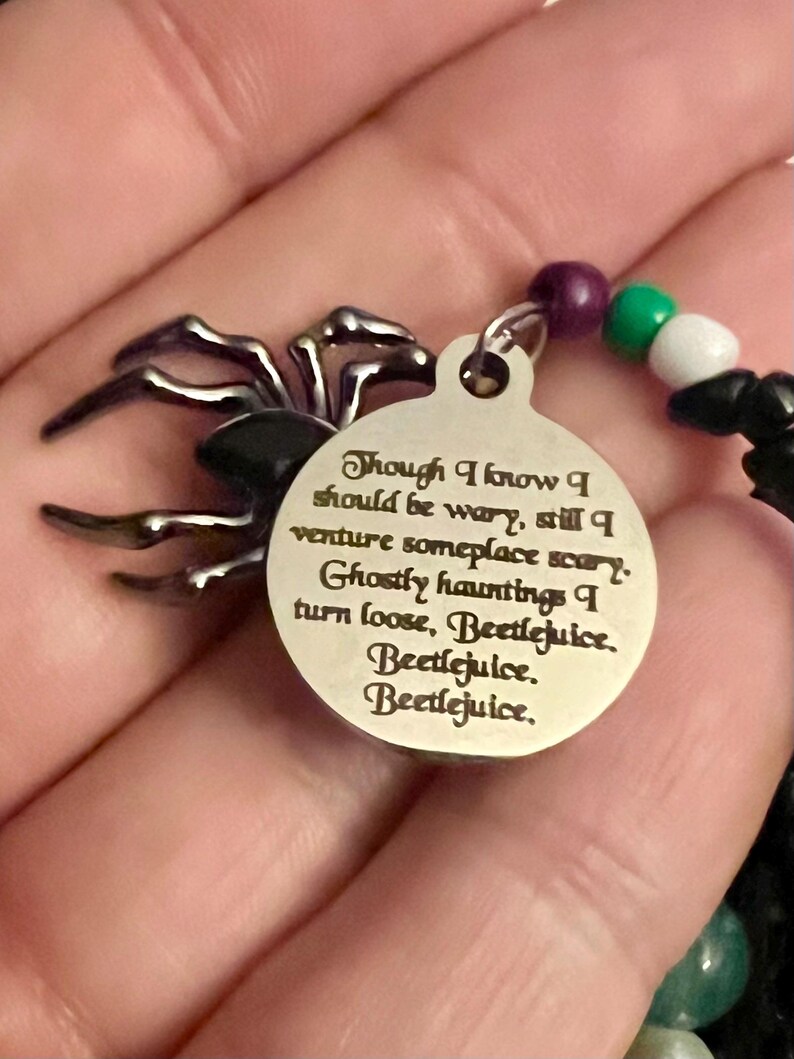 May include: A silver charm with a black spider design and a quote from the movie Beetlejuice. The quote reads: "Though I know I should be wary, still I venture someplace scary. Ghostly hauntings I turn loose, Beetlejuice. Beetlejuice. Beetlejuice."