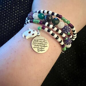 May include: A multi-strand bracelet with black and white beads, green and purple accents, and a silver charm with a ghost and the text "Though I know I should be wary, still I venture someplace scary. Ghostly hauntings I turn loose, Beetlejuice. Beetlejuice. Beetlejuice."