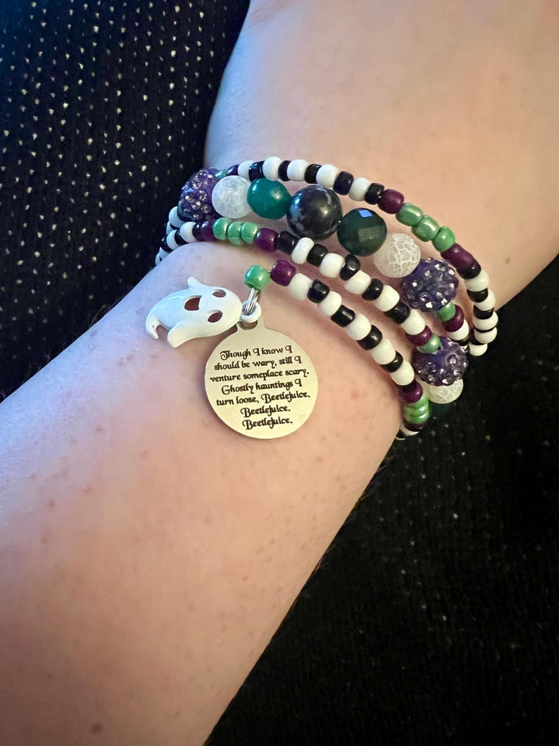 May include: A multi-strand bracelet with black and white beads, green and purple accents, and a silver charm with a ghost and the text "Though I know I should be wary, still I venture someplace scary. Ghostly hauntings I turn loose, Beetlejuice. Beetlejuice. Beetlejuice."