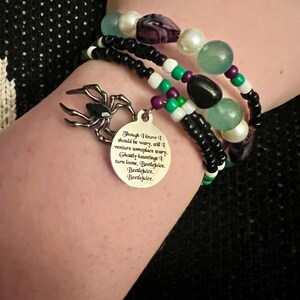 May include: A black and white beaded bracelet with a silver charm featuring a spider and a quote from the movie Beetlejuice. The quote reads: "Though I know I should be wary, still I venture someplace scary. Ghostly hauntings I turn loose, Beetlejuice. Beetlejuice, Beetlejuice."