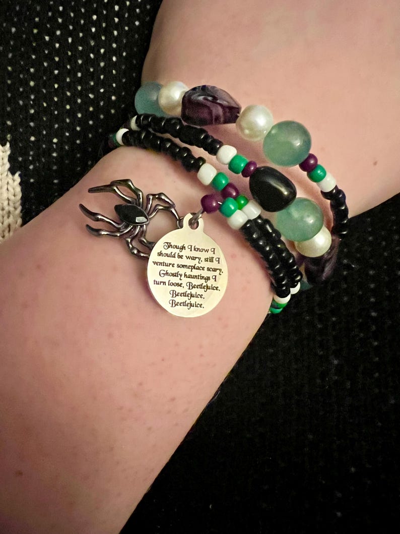 May include: A black and white beaded bracelet with a silver charm featuring a spider and a quote from the movie Beetlejuice. The quote reads: "Though I know I should be wary, still I venture someplace scary. Ghostly hauntings I turn loose, Beetlejuice. Beetlejuice, Beetlejuice."