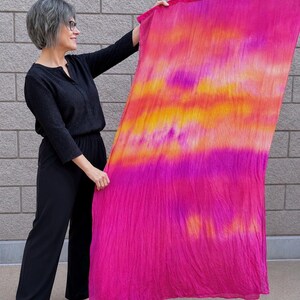 May include: A rectangular scarf featuring a vibrant tie-dye pattern with horizontal bands of bright magenta, hot pink, and golden yellow. The fabric has a lightweight, crinkled texture and is displayed fully extended to show the color gradient.