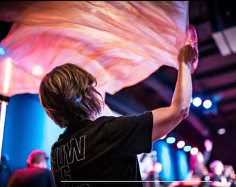 May include: A person wearing a black shirt with the text "HOW IS" printed on the back, is holding a flowing peach-colored fabric in a dimly lit room.