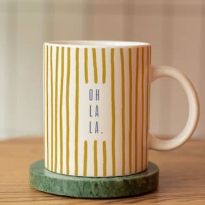 Könnte beinhalten: Eine weiße Keramiktasse mit einem gelben und weißen Streifenmuster und dem Text "OH LA LA." Die Tasse steht auf einem grünen Marmoruntersetzer.