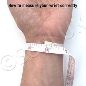 May include: A white measuring tape wrapped around a wrist to show how to measure wrist size correctly. The tape reads 5, 6, 7, 20, 21, 22, 23, and 24 inches.