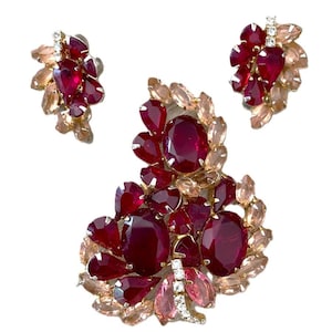 May include: A jewelry set consisting of a large brooch and two matching earrings. The pieces feature deep red oval and pear-shaped faceted stones surrounded by smaller, pale pink marquise-cut stones set in a gold-toned metal frame.
