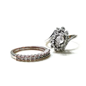 May include: Two silver rings with clear gemstones. One ring features a band of small stones, while the other has a cluster of stones in a floral design. The rings are set against a white background.
