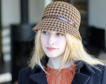 May include: A woman wearing a brown and black houndstooth patterned hat with a brown leather band. She is wearing a black jacket and a rust colored blouse.