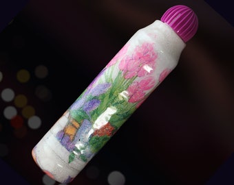 May include: A white glitter marker with a pink floral design and a purple cap. The flowers are pink, purple, and green.