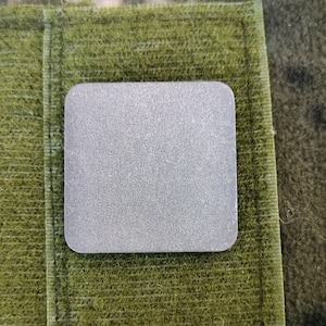 以下が含まれることがあります： 表面がテクスチャ加工された、小さな正方形の銀色の金属プレート。プレートは、緑色の生地に「CALTI TITANIUM EDC」という文字が印刷された状態で取り付けられています。