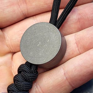 Puede incluir: Una cuenta cilíndrica de titanio gris con una superficie texturizada, unida a un cordón trenzado negro. La cuenta se sostiene en una mano. El texto "CALTI TITANIUM" es visible en la esquina inferior izquierda.
