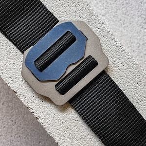 Puede incluir: Una correa de nylon negra con una hebilla de metal plateada y azul. La hebilla es rectangular con bordes redondeados y una superficie lisa.