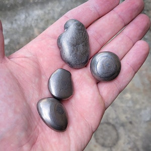 Könnte beinhalten: Vier glatte, graue Titan-Steine in verschiedenen Formen und Größen, die in einer Hand gehalten werden. Die Steine sind glatt und haben einen metallischen Glanz. Der Text "CALTI TITANIUM EDC" ist in der linken unteren Ecke des Bildes sichtbar.