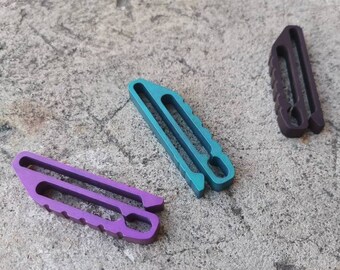 Pode incluir: Três pequenos clipes de metal em tons de roxo, verde-azulado e roxo escuro estão dispostos sobre uma superfície cinza texturizada. Cada clipe tem um design único com uma abertura ranhurada e uma borda estriada, sugerindo um propósito funcional ou decorativo.