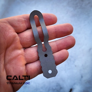 Pode incluir: Uma ferramenta EDC de titânio cinza com um design de buraco de fechadura, segurada em uma mão. A ferramenta tem um topo arredondado com uma abertura longa e estreita e uma borda inferior texturizada. O texto "CALTI TITANIUM EDC" é impresso na parte inferior.