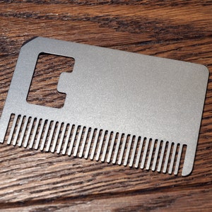 Puede incluir: Un peine de metal plateado con un abridor de botellas. El peine tiene una fila de dientes y una gran abertura rectangular para el abridor de botellas.