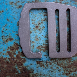 Op de afbeelding: Een zilveren metalen sleutelhanger met een D-vormige ring en drie sleuven. De sleutelhanger ligt op een blauw en bruin getextureerd oppervlak.