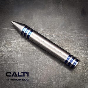May include: A silver-colored titanium EDC pen with blue accents. The pen has a pointed tip and a patterned design on the body. The text "CALTI TITANIUM EDC" is displayed below the pen.