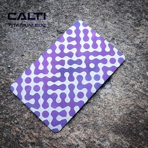 May include: A rectangular titanium EDC card with a purple and white maze-like pattern. The card has rounded corners and the text "CALTI TITANIUM EDC" is visible at the top. The card is set against a textured gray background.