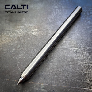 May include: A silver-colored titanium EDC pen with a pointed tip. The pen is cylindrical and has the text "CALTI TITANIUM EDC" in the upper left corner. The pen is lying on a dark, textured surface.