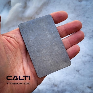 Puede incluir: Una tarjeta EDC rectangular de titanio gris, con esquinas redondeadas, se sostiene en una mano. La tarjeta tiene una superficie texturizada. El texto "CALTI TITANIUM EDC" está impreso en negro en la parte inferior de la imagen.