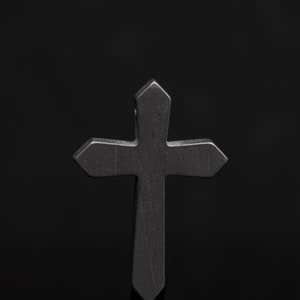 Puede incluir: Una cruz de metal negro con una parte superior puntiaguda y bordes redondeados. La cruz está de pie sobre una superficie negra.