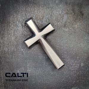 Op de afbeelding: Een zilverkleurige kruisvormige hanger met een rechthoekige balk en schuine armen. De tekst "CALTI TITANIUM EDC" is eronder in zwart gedrukt. Het kruis is gemaakt van titanium.