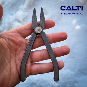 May include: A dark gray multi-tool pliers with the text "CALTI TITANIUM EDC". The pliers are open, revealing the cutting edges. The tool is held in a hand against a snowy background.