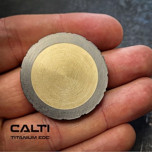 Op de afbeelding: Een ronde, tweekleurige metalen munt met een gouden midden en een zilveren buitenring. Het gouden midden heeft een spiraalpatroon. De munt wordt in een hand gehouden. De tekst "CALTI TITANIUM EDC" is in het zwart gedrukt.